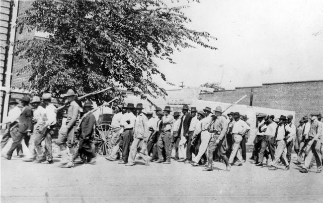 Nacionālās gvardes karaspēka grupa, nesot šautenes ar piestiprinātām bajonetēm, pavada neapbruņotus melnādainos vīriešus uz aizturēšanas centru pēc Talsas rases slaktiņa Talsā, Oklahomas štatā, 1921. gada jūnijā.