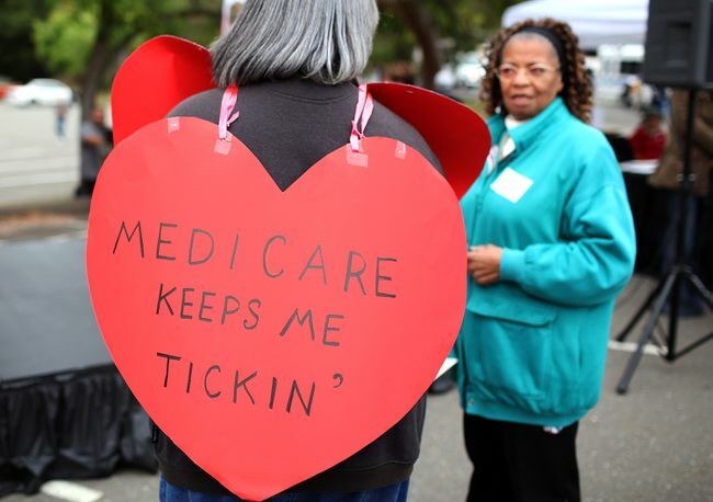 Sieviete ar sirds formas izkārtni ar uzrakstu “Medicare Keeps Me Ticking”