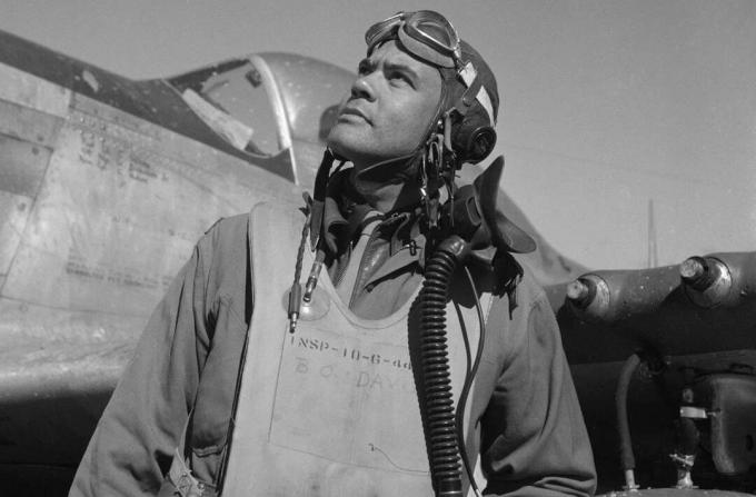 Bendžamins O. Deiviss lidojuma uzvalkā un ķiverē, kas stāv pretī iznīcinātājam P-51 Mustang.