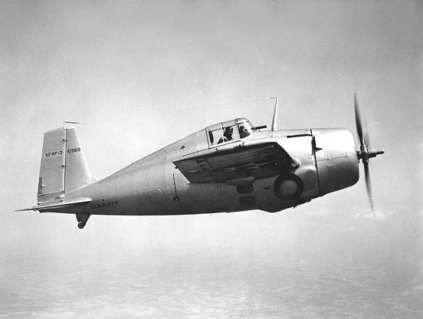 Grumman XF4F-3 Wildcat lido pa kreisi uz labo pusi, sudraba alumīnija apdare, pilots skata uz āru.
